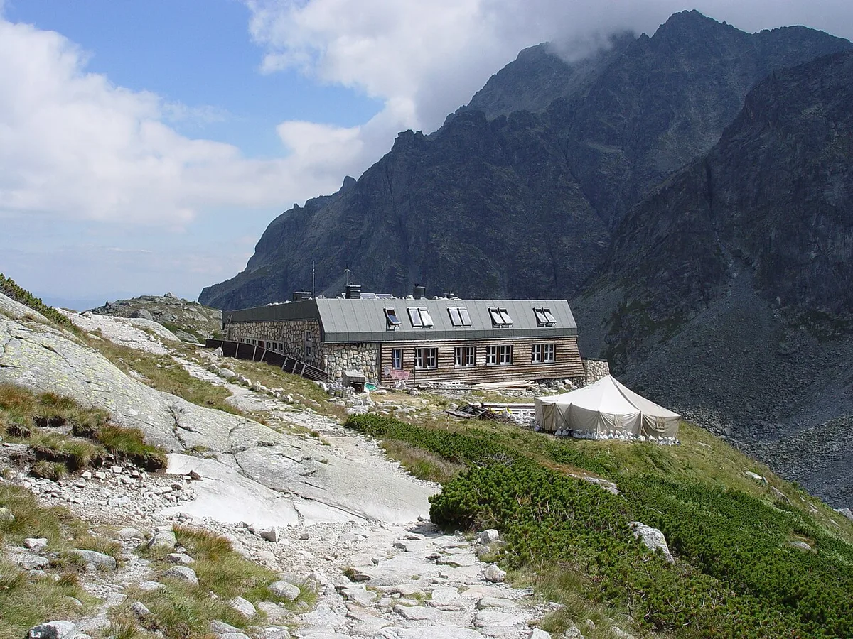 Een houten berghut in de Tatra tegen een steenhelling, met een groepje wandelaars aan de lange tafels ervoor.