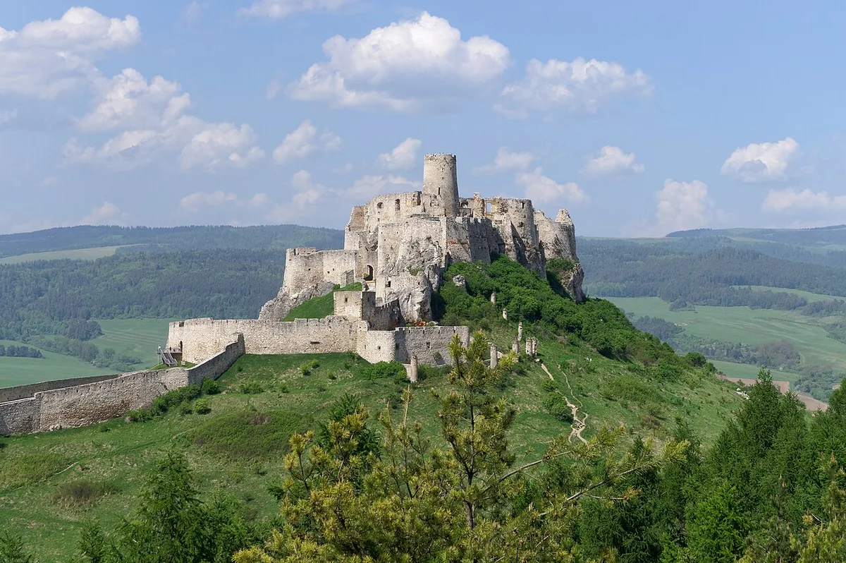 De uitgestrekte witte muren en de ronde donjon van Spišský hrad op een groene heuvel onder een wolkenlucht.